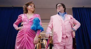 The Beanie Bubble: Elizabeth Banks as Robbie and Zach Galifianakis as Ty, shot from a low angle, both wearing pink outfits and looking at each other with a smirk. He is leaning on a pink cane, and she has a blue stuffed plush Himalayan cat under one arm