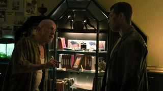 An older man wearing a white vest and plaid shirt is having a heated conversation with a younger, taller man standing in front of him. In the background you can see a lit bookshelf and a fishtank.