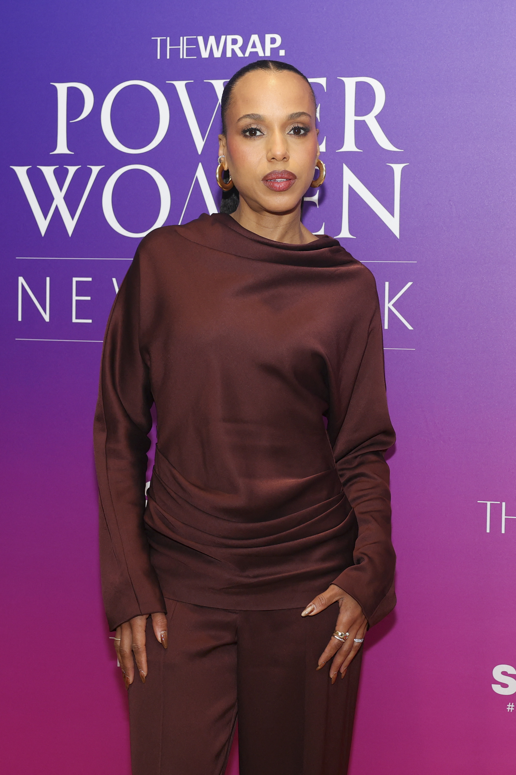 a black woman wearing a maroon pants set and posing in front of a pink and purple background