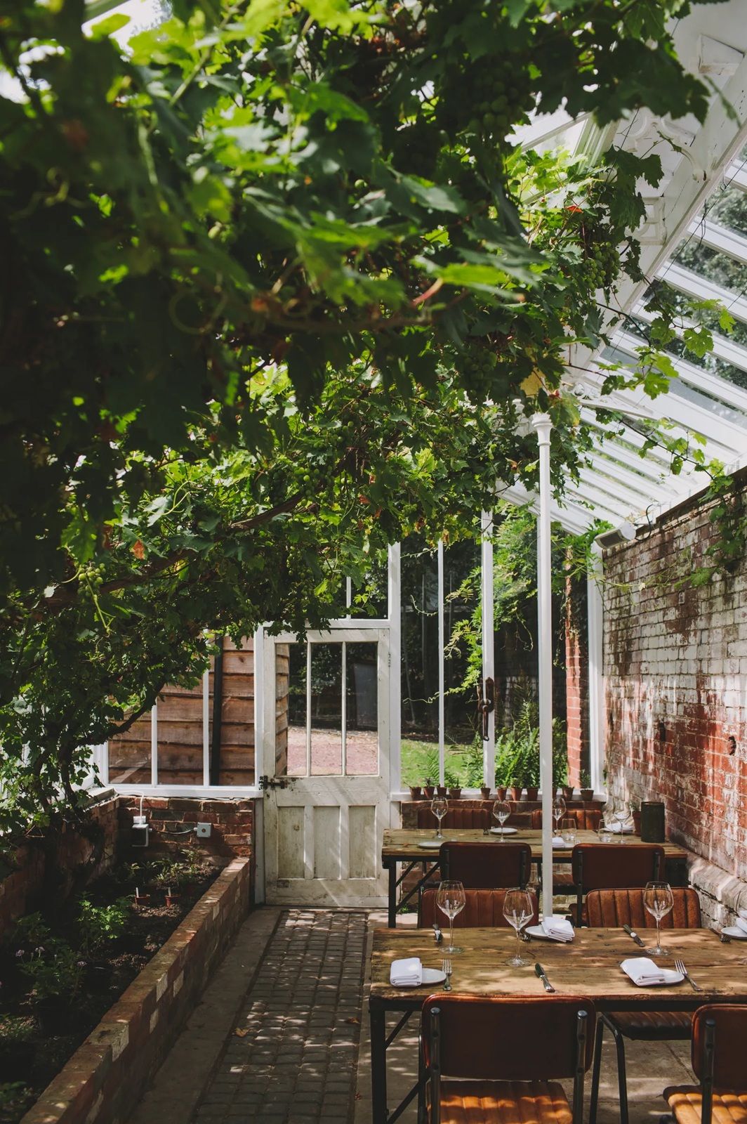 A walled garden has been transformed into an intimate, rustic dining area, with wood and leather furniture and minimal decor contrasting with lush vegetation and the red of bricks.