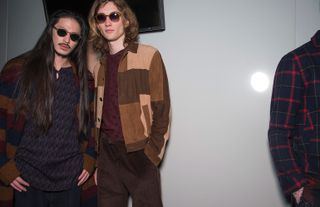 Two male models wearing looks from Oliver Spencer's collection. One model is wearing a dark blue patterned top and brown, blue and maroon cardigan. And the model next to him is wearing a patterned maroon coloured top, brown trousers and a jacket with sections in different shades of brown