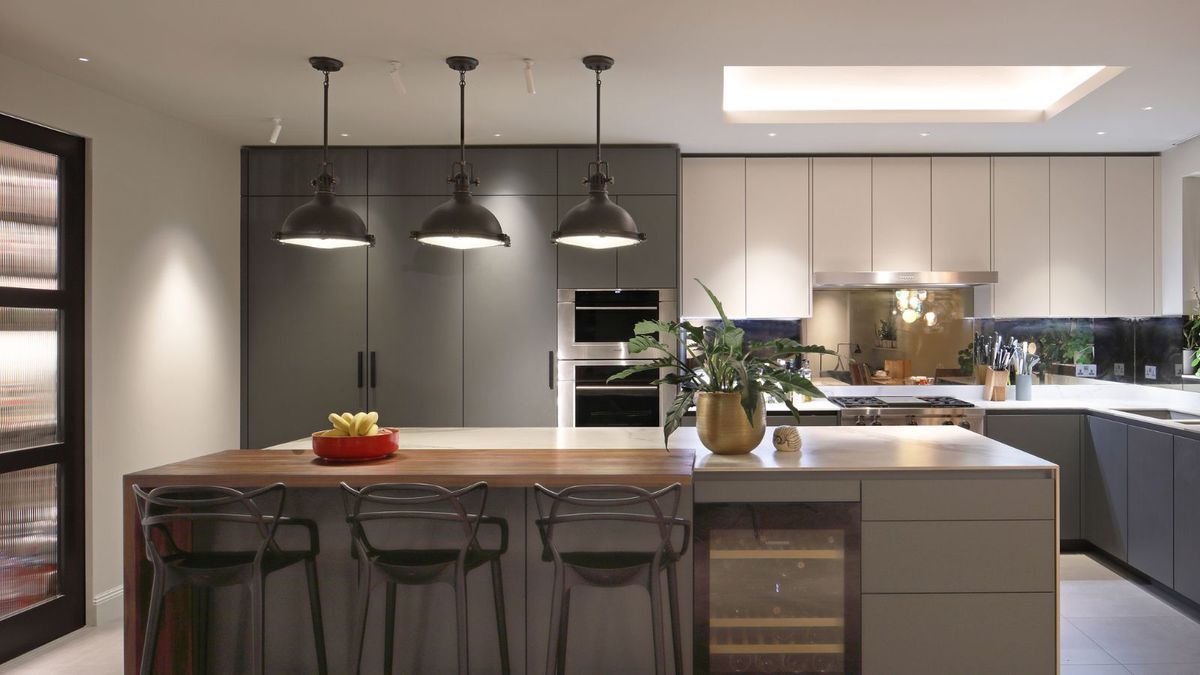 A modern kitchen featuring a large island with a wooden countertop and three black bar stools. Overhead, three industrial-style pendant lights provide focused illumination. The cabinetry combines dark green and white finishes, with integrated appliances and a mirrored backsplash. A gold planter and a bowl of bananas decorate the island.