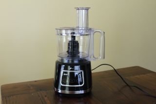 The black Oster 10-Cup Food Processor with Easy-Touch against a cream wall
