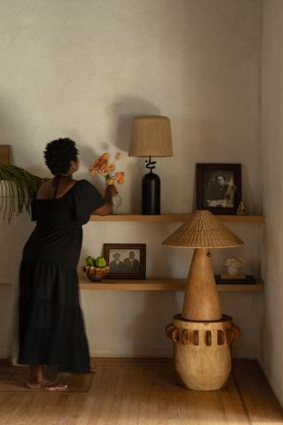 A woman in a black maxi dress fixes some flowers in a vase positioned on a wooden shelf alongside a rattan lamp, a framed photographed. Below it is another shelf, also decorated with plants and framed photographs. To her right, a rattan and wood standing lamp.