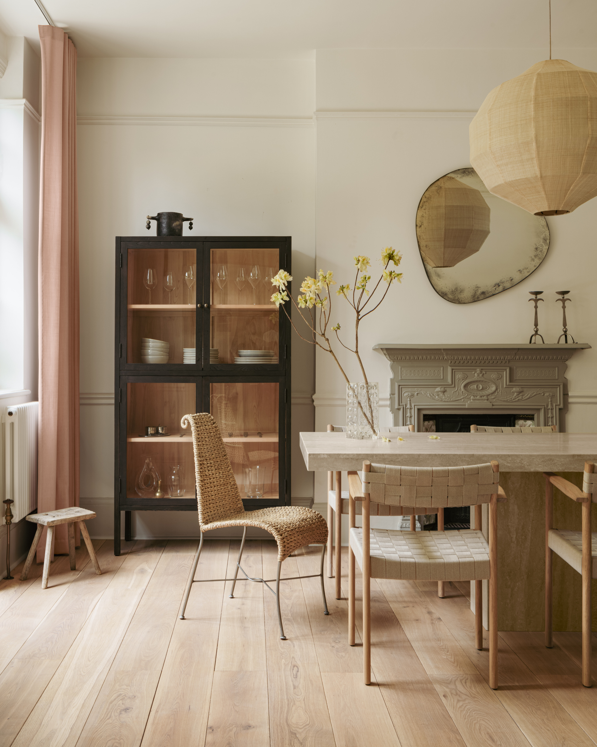 neutral dining room with neutral walls, stone table, mis-matched dining chairs, timber floorboards, large round pendant light, artwork over original fireplace, hosting cupboard with glass doors filled with crockery, small stool next to it, and pink curtains