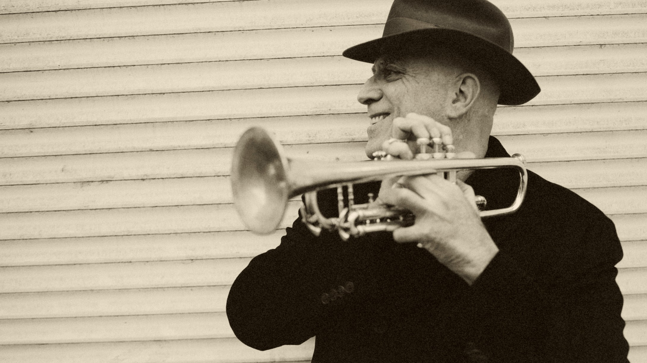 An older man in a hat smiles as he holds a trumpet. The image is mostly in gray-scale and the vibe is very cool.