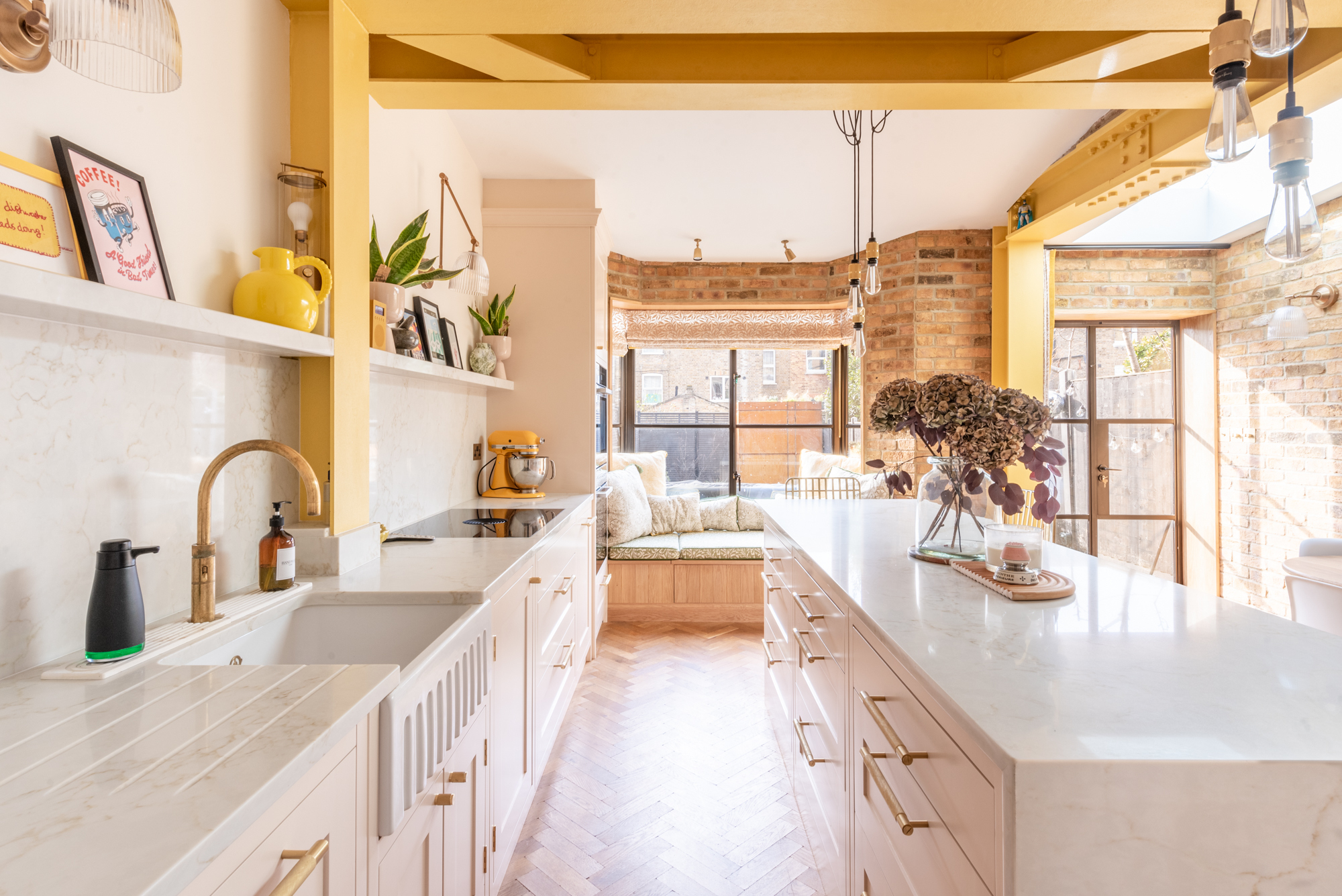 white kitchen with brass handles, yellow beams, exposed brickwork on the walls, and window seat and herringbone floors