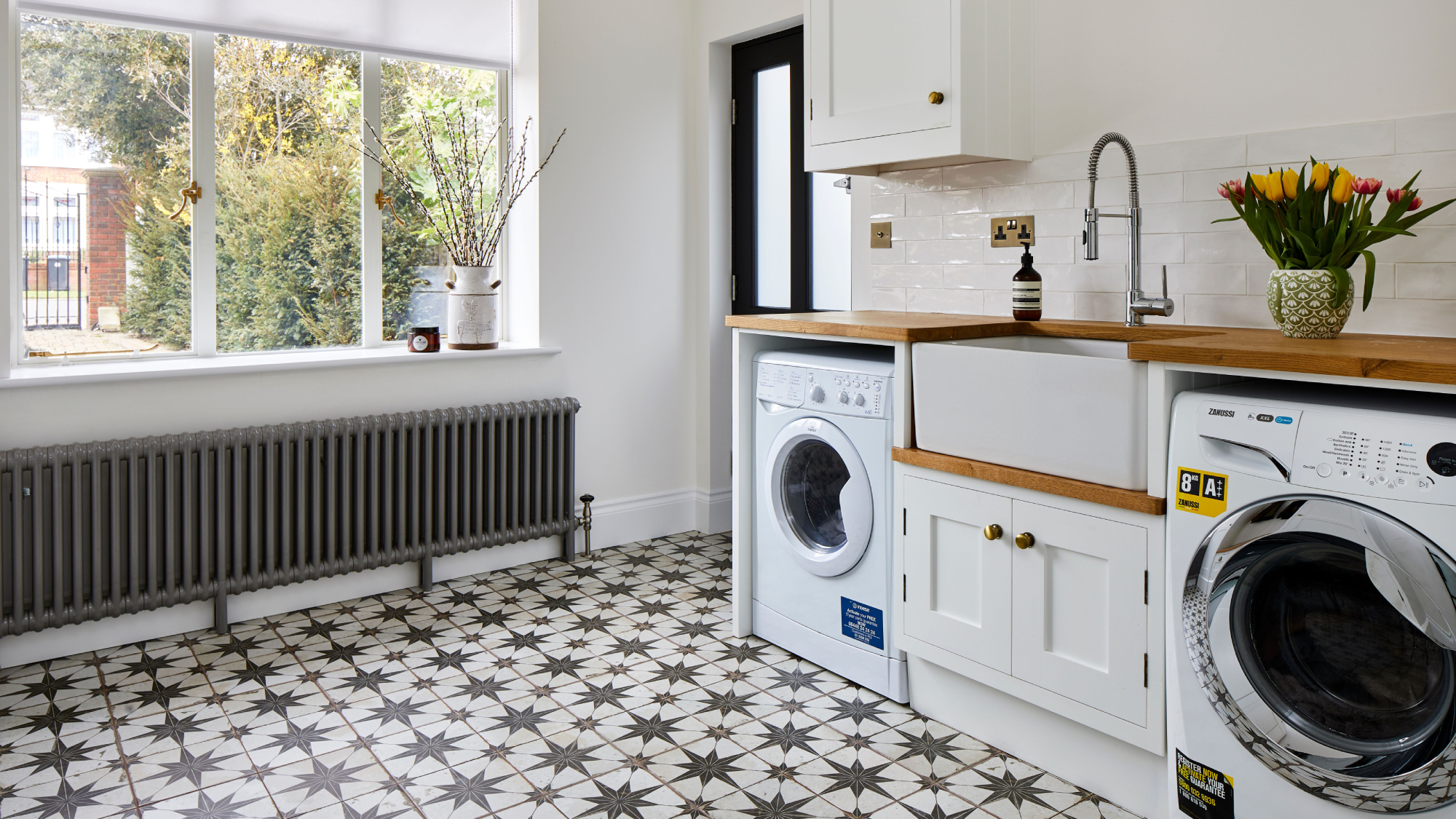 utility room with black and white star tiled floor, large window, grey radiator, appliances, butler sink, wooden worktop and white cabinets