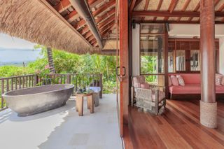 A tropical, luxury hut features wood and rattan interiors, an outdoor bathtub, and views of the tropical nature all around it.