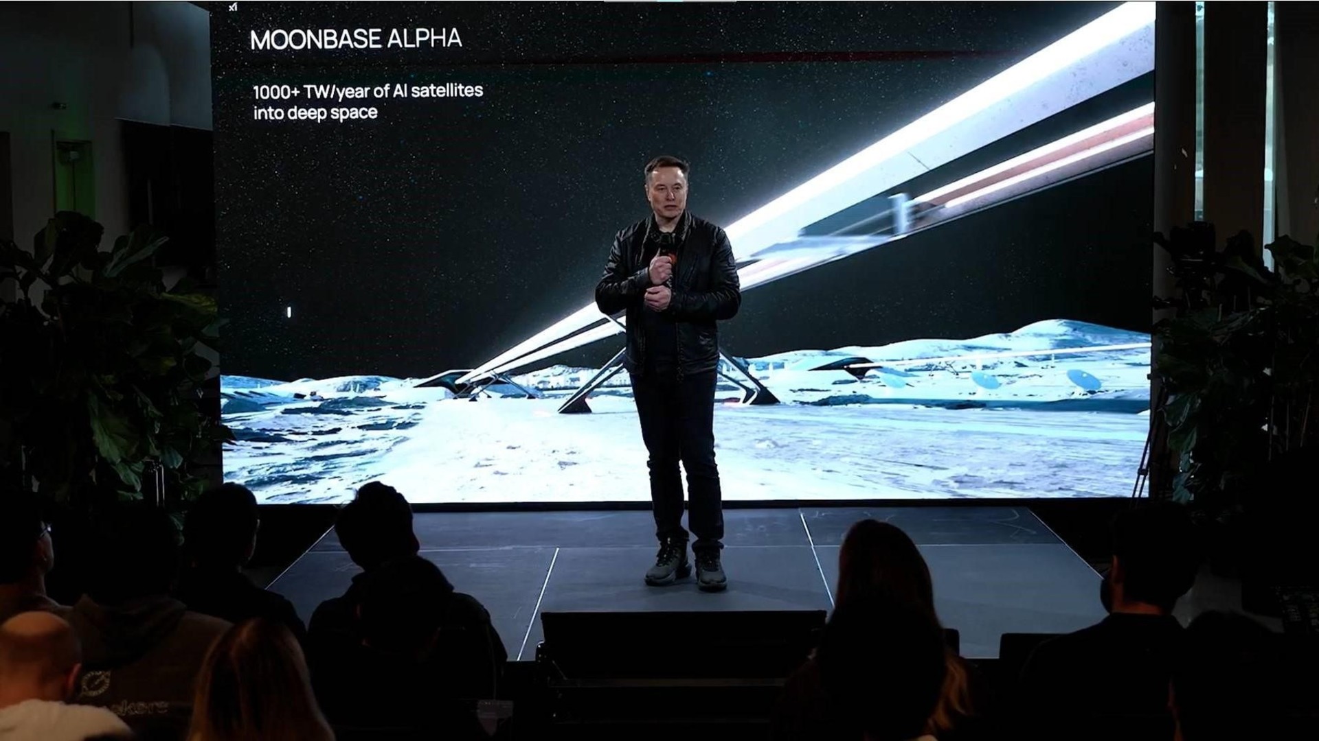 Um homem vestindo uma camiseta preta e jeans pretos fala em um microfone no palco, em frente a uma ilustração de uma estrutura semelhante a uma ferrovia na lua