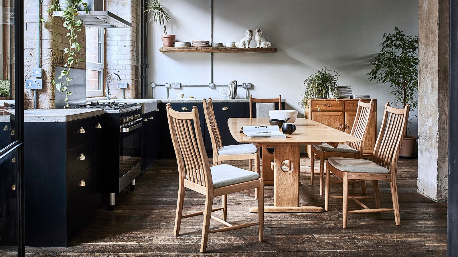 A kitchen diner with black kitchen cabinets and a large wooden dining table, surrounded by five matching wooden chairs. 