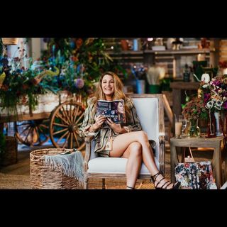 Blake Lively sits in a chair holding a copy of the book It Ends With Us while wearing a shirt she borrowed from husband Ryan Reynolds along with strappy Versace heels