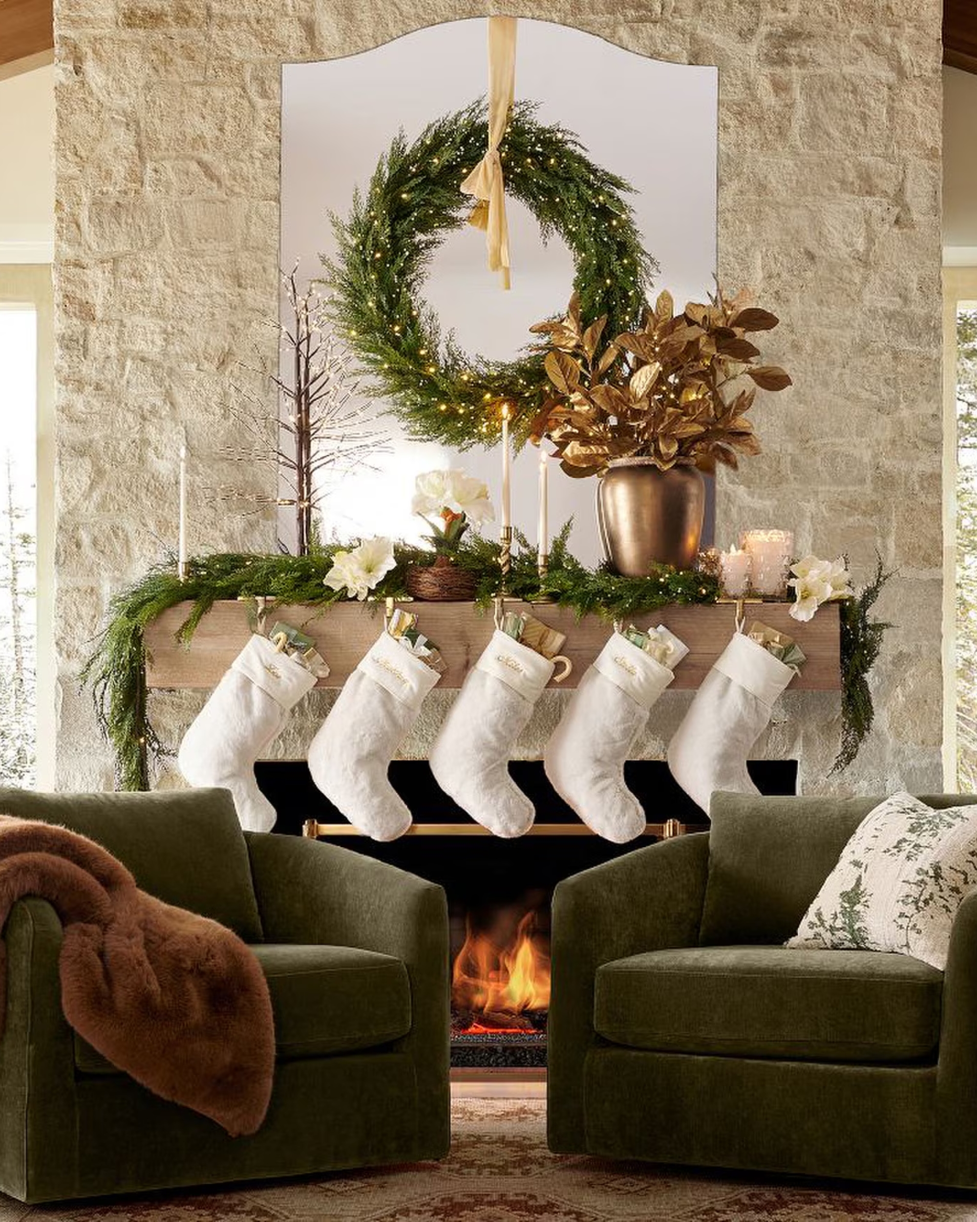Image of a living room with a large stone fireplace that is decorated for Christmas &amp;mdash; there is a wreath, garland, and five white stockings. Two green accent chairs are in front of the fireplace.