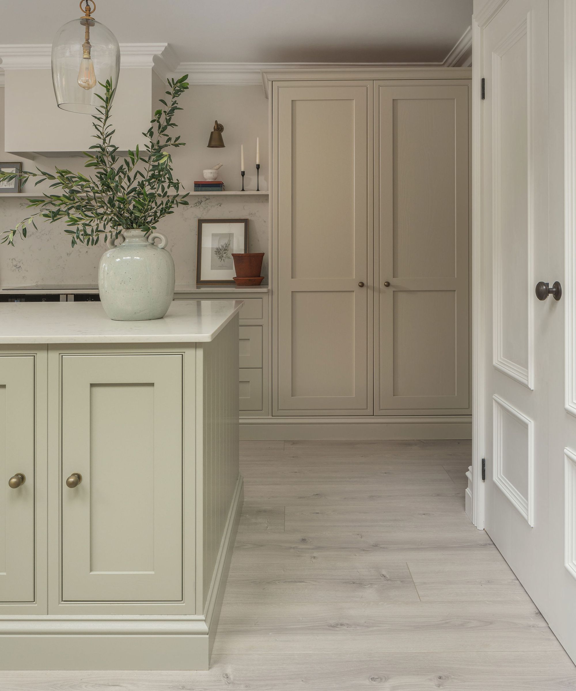 Calm neutral kitchen with soft sage and beige cabinetry, pale wood floors, stone counters, and subtle brass details creating a serene, timeless space.