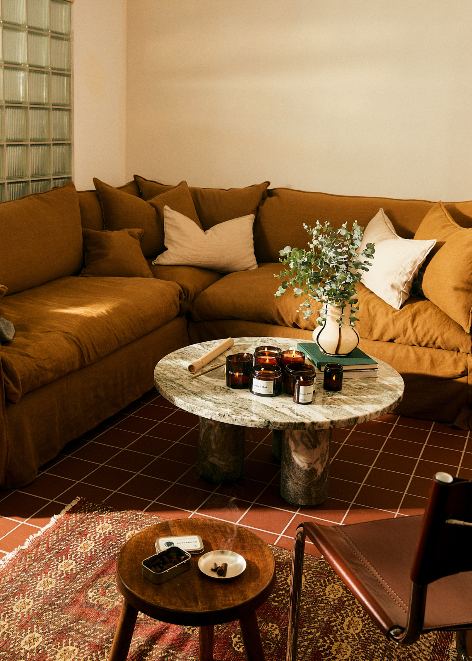 A cozy living room with a stack of pillows by a marble coffee table with a set of candles, a vase of eucalyptus and some books