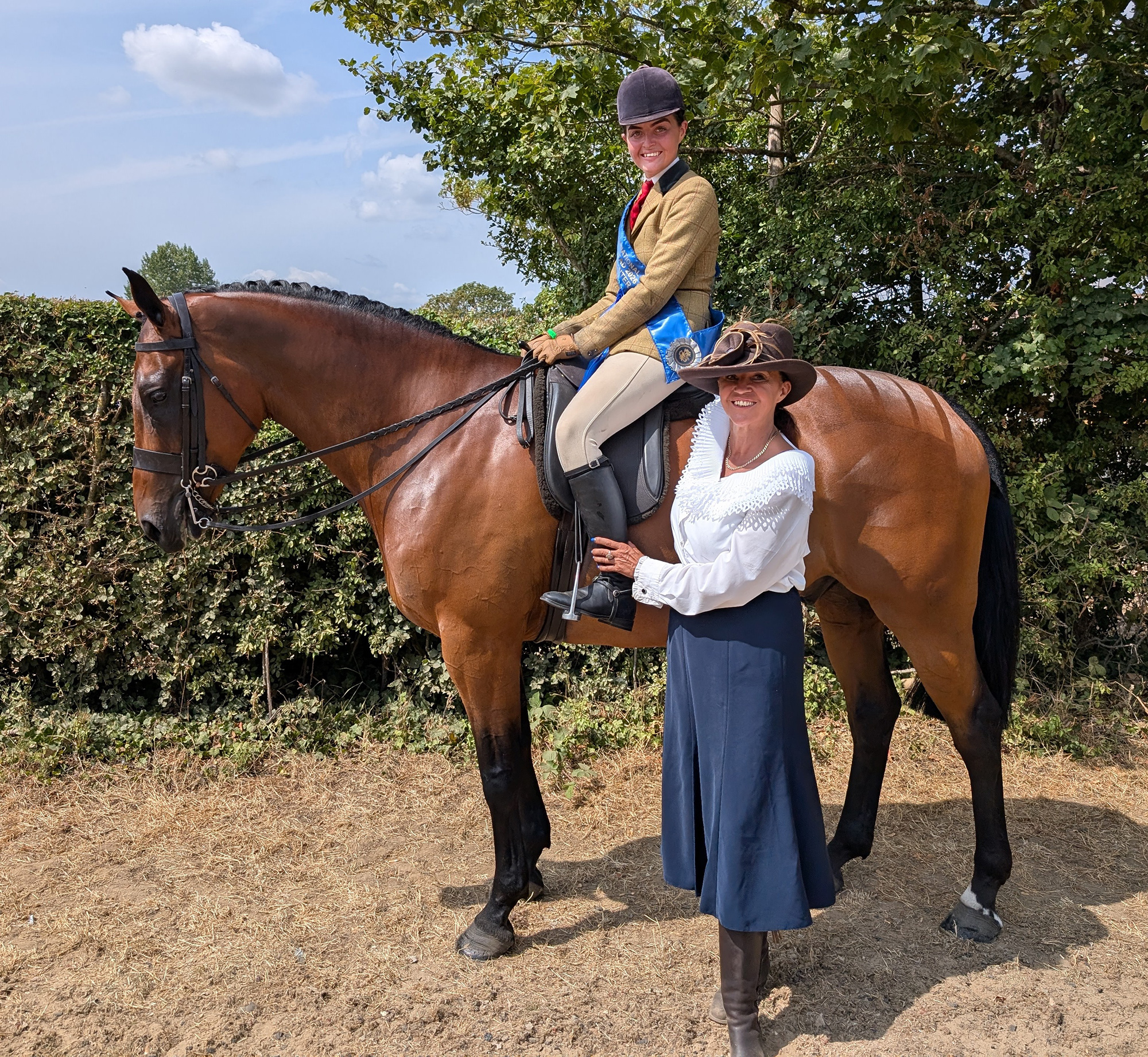 A bay small hunter with rider wearing blue winner's sash with mum wearing blue skirt, white blouse and brown hat standing alongside