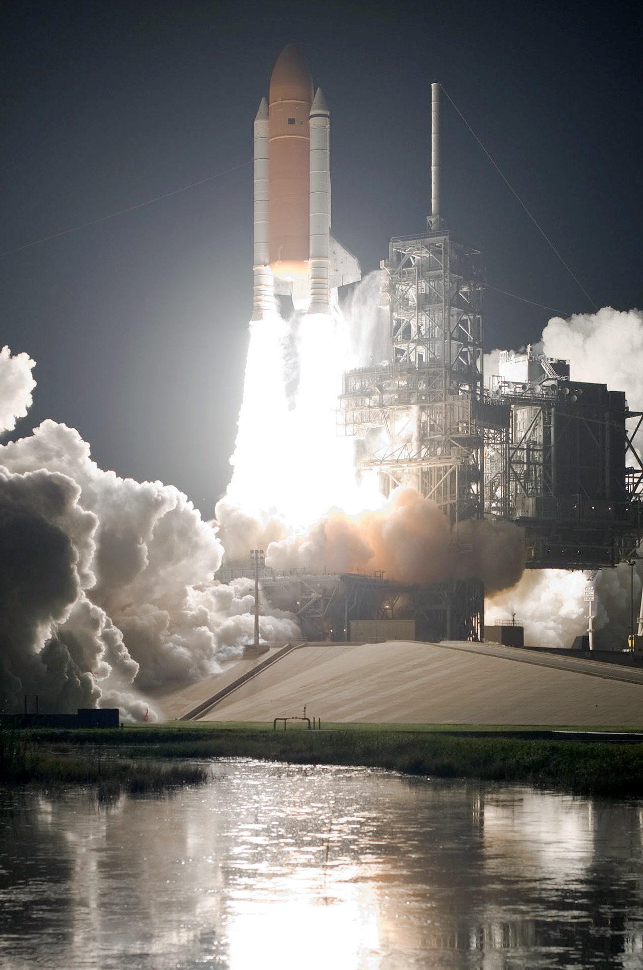 space shuttle lifting off at night, with a reflection in the water in front