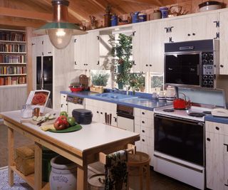 Interior view of a large blue and white kitchen with a a center counter space laid with apples and a cutting board, circa 1980.