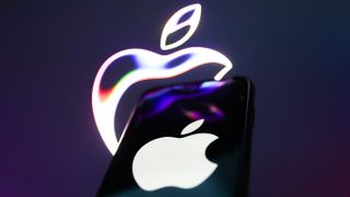 An iPhone bearing the Apple logo on a dark screen, with purple light reflecting on it from a large stylised Apple logo behind it, which is lit against a purple background.