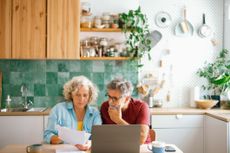 A mature couple sits at a kitchen table with a laptop and papers.