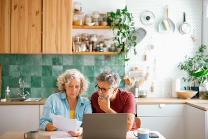 A mature couple sits at a kitchen table with a laptop and papers.