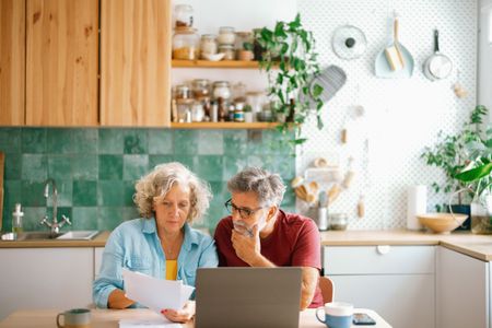 A mature couple sits at a kitchen table with a laptop and papers.