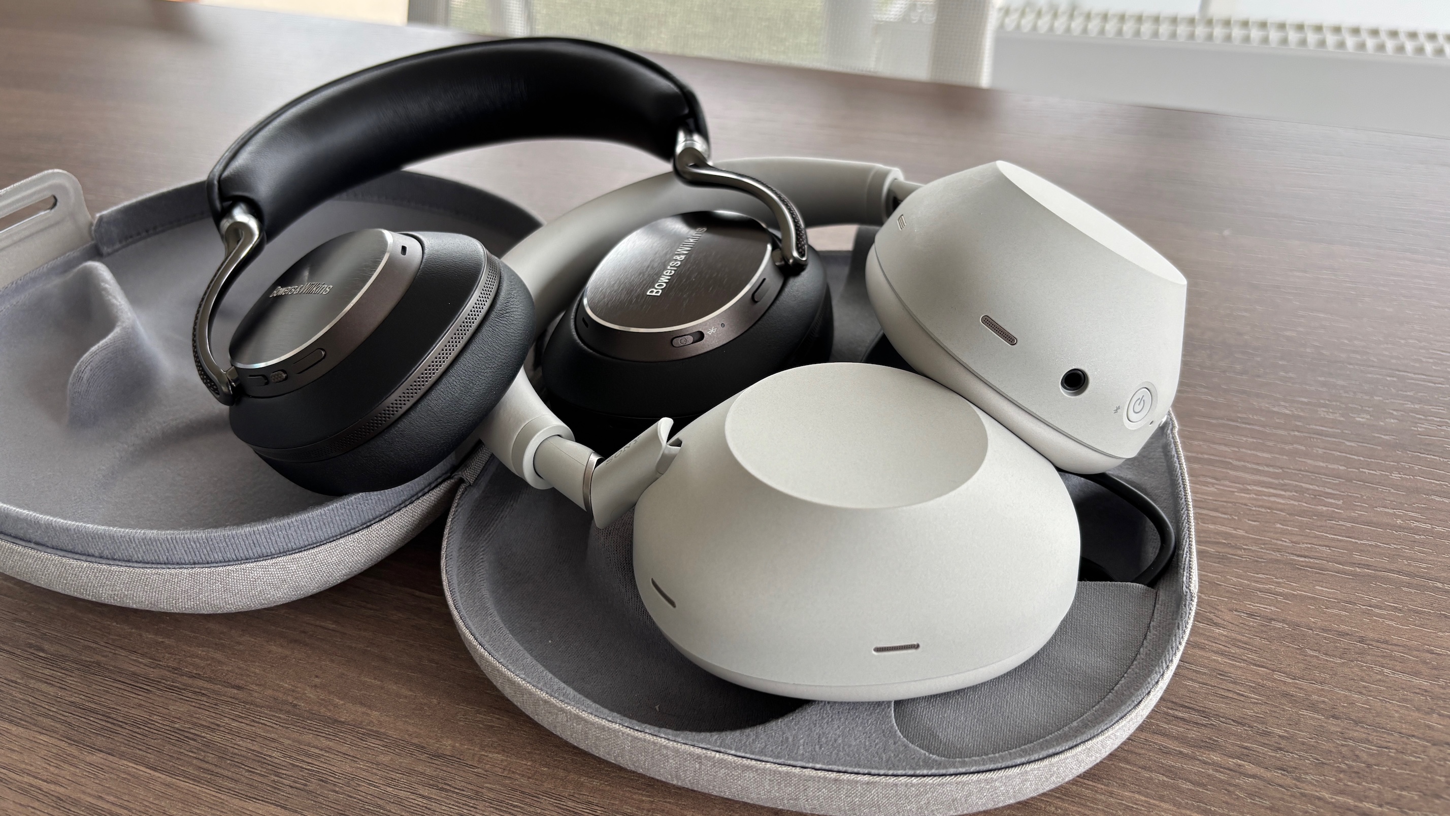 Bowers &amp;amp; Wilkins Px8 S2 and Sony WH-1000XM6 headphones in their carry cases on a wooden table.