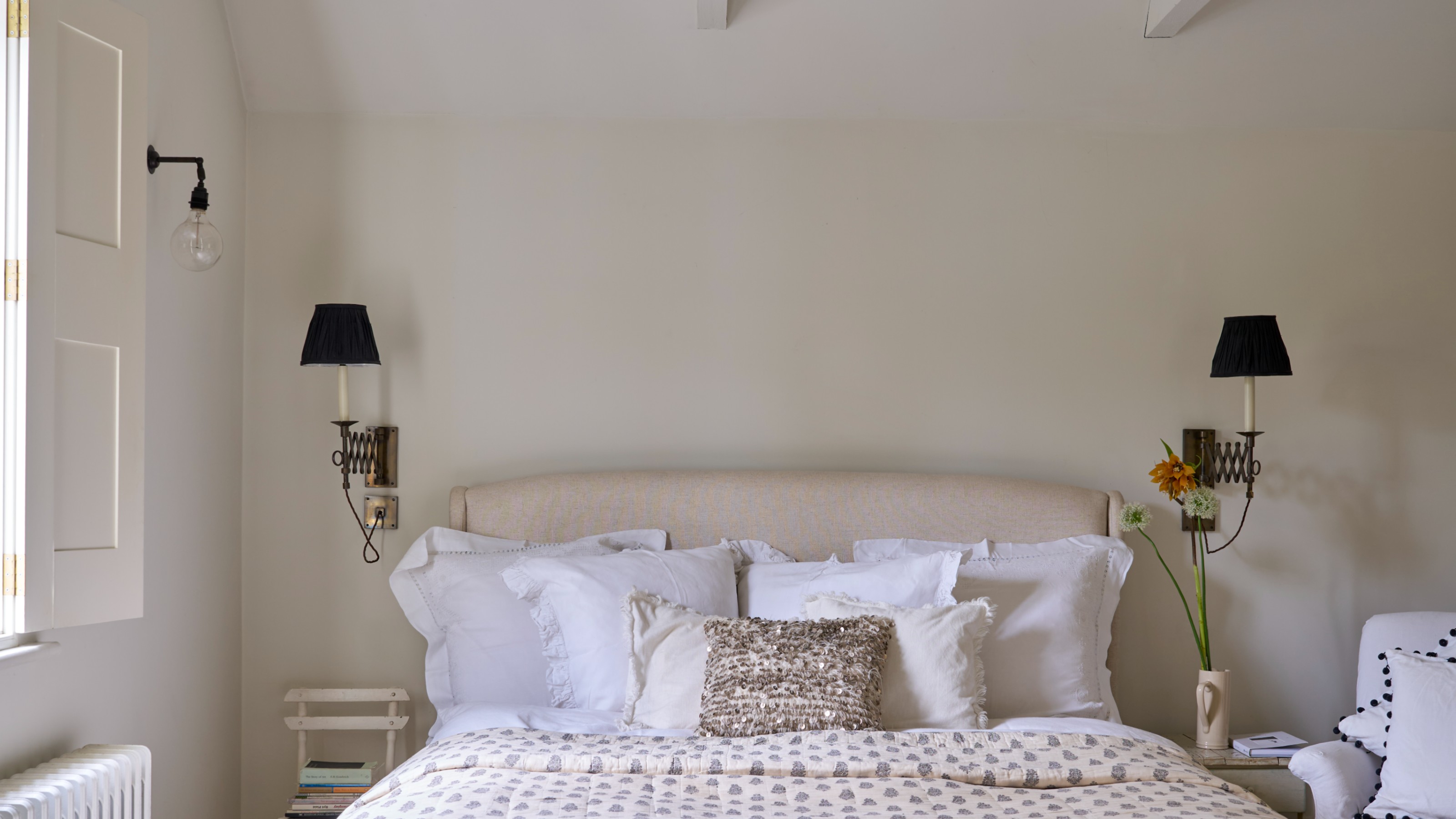 A bedroom painted in Farrow &amp; Ball's Wimborne White shade with an upholstered cream headboard and white bed linen