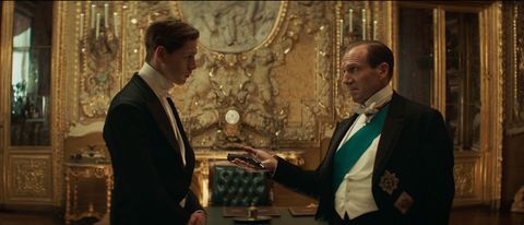 Harris Dickinson is offered a gun by Ralph Fiennes in a well dressed room in The King's Man.
