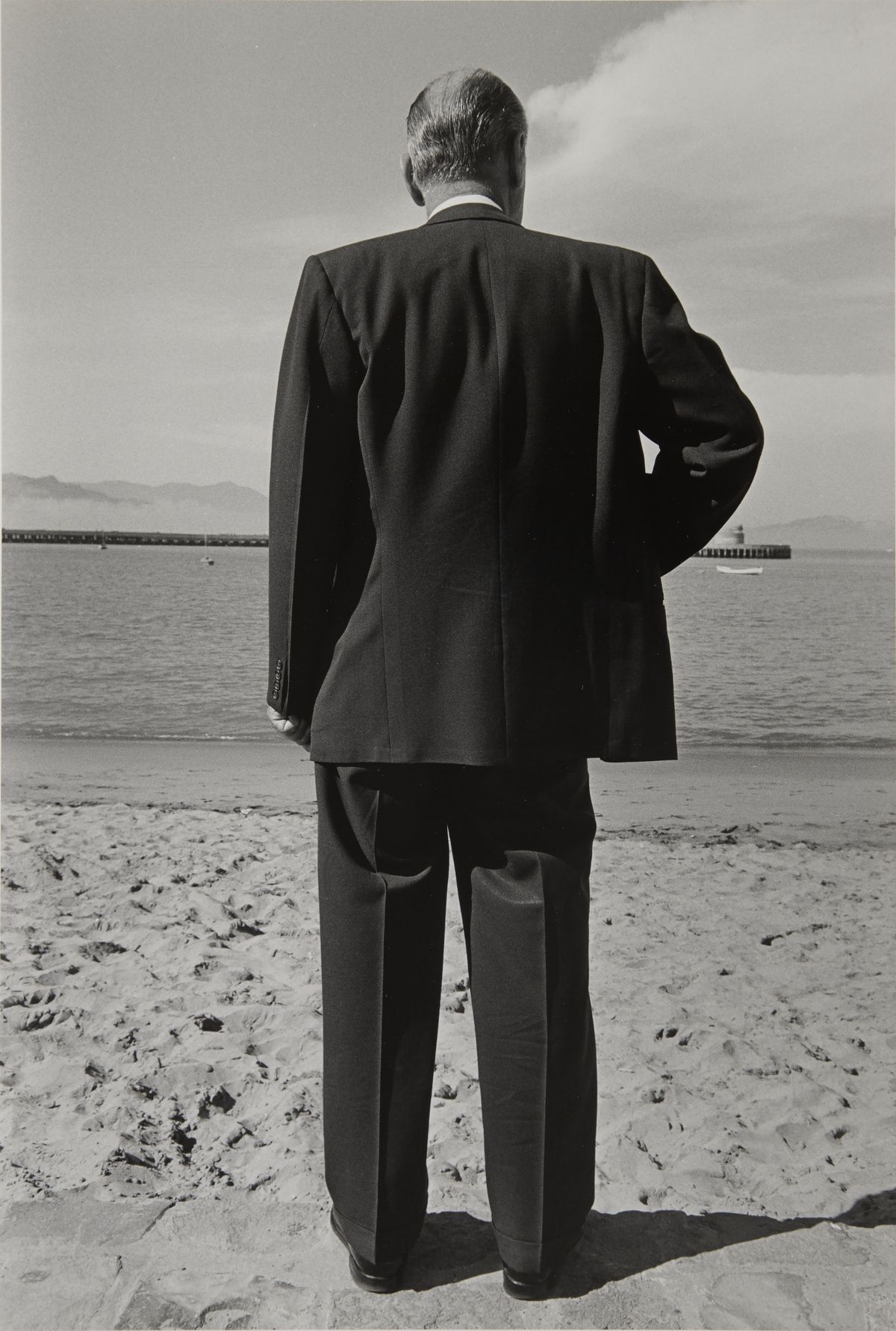 A black and white photograph of a man in a dark suit, seen from behind, standing on a beach and facing out toward the water.