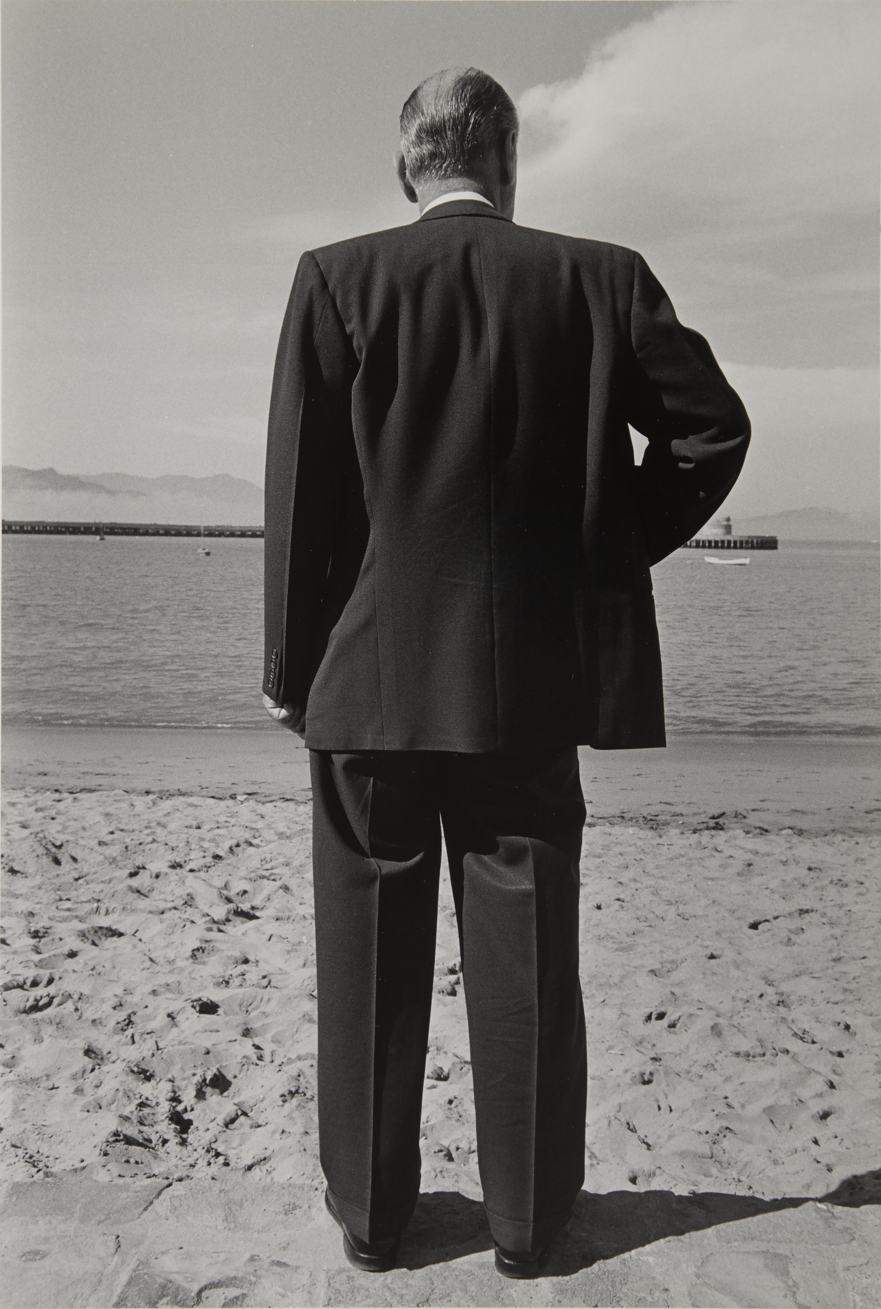 A black and white photograph of a man in a dark suit, seen from behind, standing on a beach and facing out toward the water.