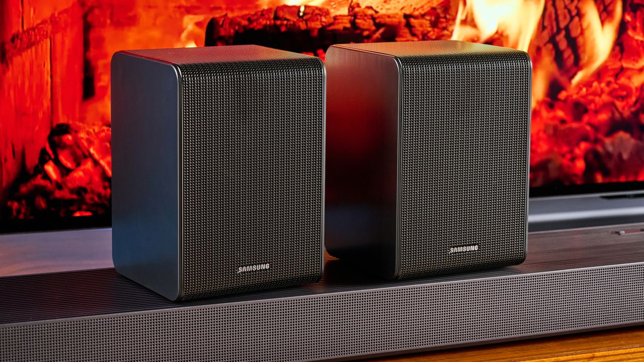 the samsung qs750f soundbar with two rear surround speakers and subwoofer photographed against a blue background and a TV