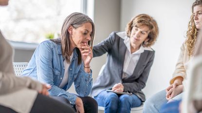 A woman places her hand on the shoulder of another woman to comfort her in a waiting room