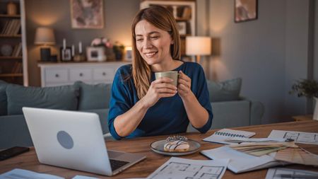 Women in a cosy living room, holding a hot drink and surrounded by house plans and colour swatches, talking to an expert on her laptop