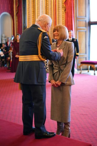 Anna Wintour wearing a gray skirt suit smiling at King Charles, who is standing on a red platform and pinning a ribbon on her jacket at Buckingham Palace.