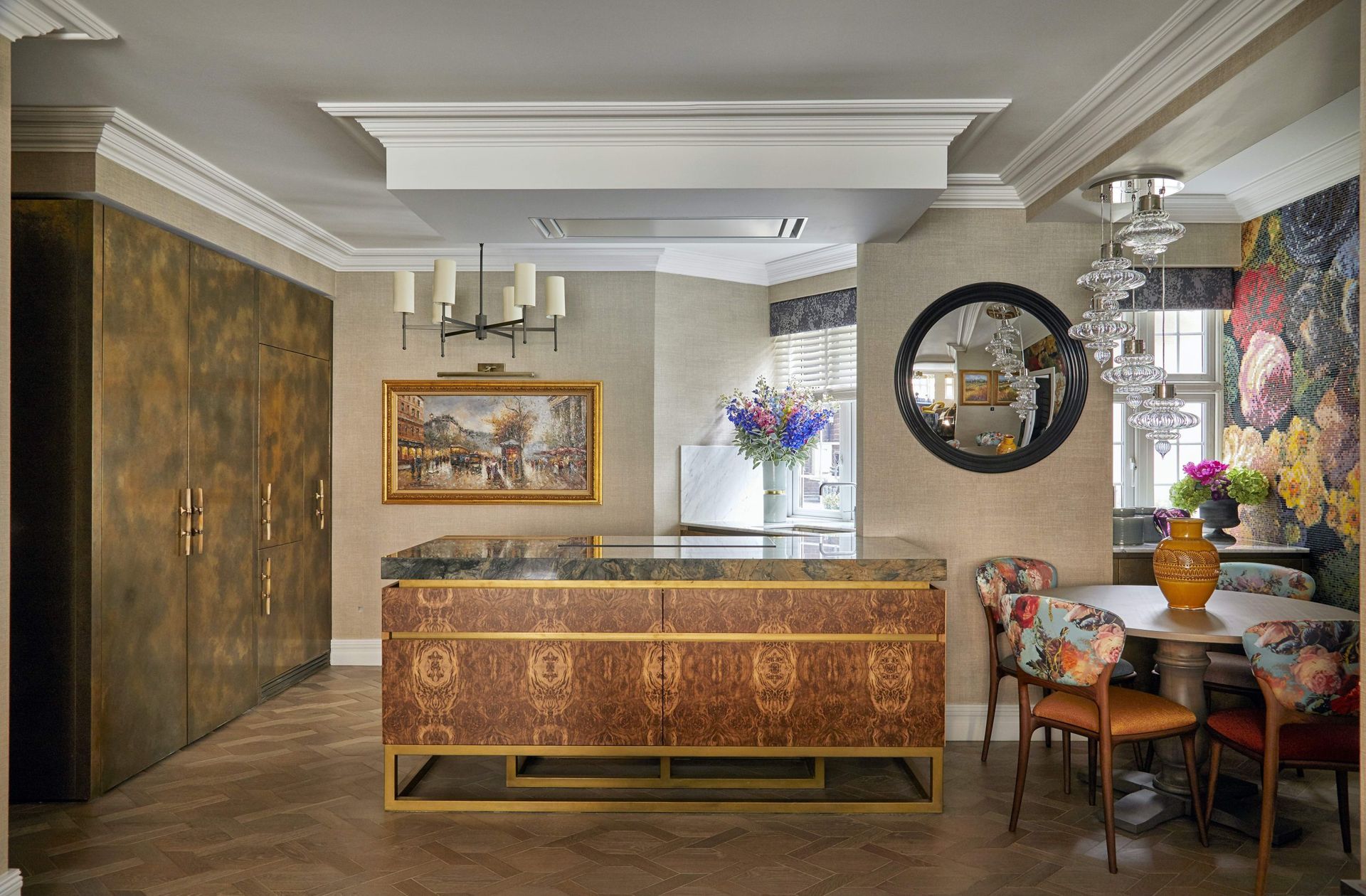 A kitchen, dining and living space with brown floor to ceiling cabinets, a red and gold island, and a circular table with floral chairs