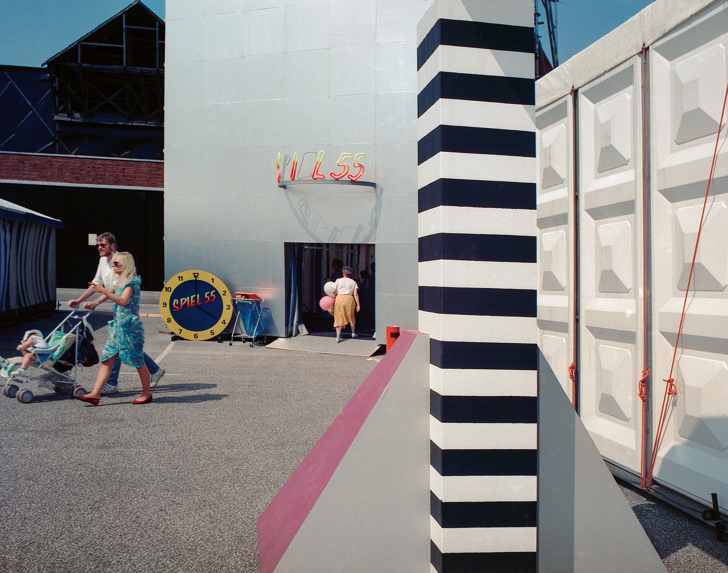 Outside the "Spiel 55" fairground attraction in 1989 Cologne, a couple push a pram past a giant novelty clock, set against a striped concrete column and shipping-container-style wall.
