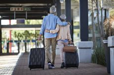 An older couple walking together on public transport station. They are walking away from the camera, pulling rolling suitcases and bag behind them.