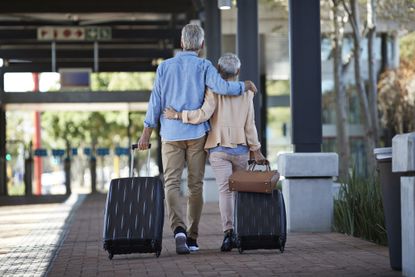 An older couple walking together on public transport station. They are walking away from the camera, pulling rolling suitcases and bag behind them.
