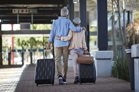 An older couple walking together on public transport station. They are walking away from the camera, pulling rolling suitcases and bag behind them.
