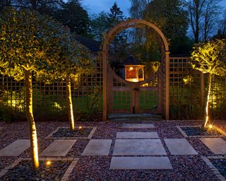 Garden feature Arch at night with holly trees lit and gates leading into lawn area with illuminated Gazebo