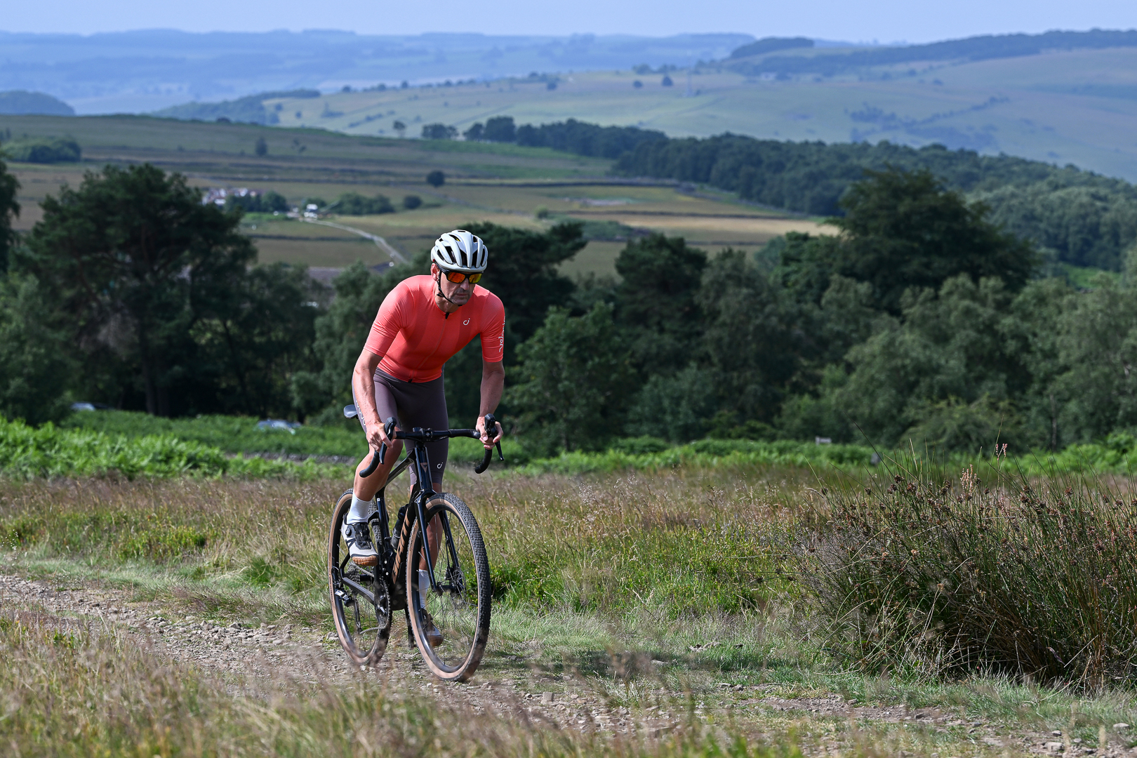 Man in an orange jersey and brown shorts riding out of the saddle towards the camera on a trail lined with grass