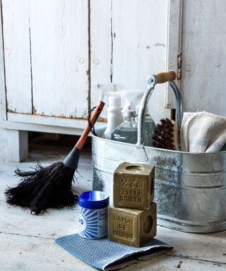 Rustic, distressed utility room, wooden floorboards, metal bucket containing cleaning products and brushes, feather duster, blocks of soap and polish.