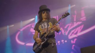 Slash wears a top hat and plays a Gibson SG live with Guns N' Roses, with pink and blue lights in the background.
