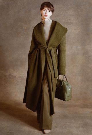 One of the best jacket coat brands, Staud, is shown in an image of a model wearing a green coat over a tan turtleneck, green pants, and tan boots