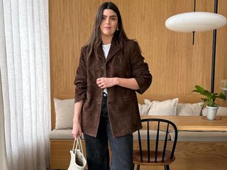 British fashion influencer Anna Newton poses in her modern London kitchen with wood panelling wearing a brown suede jacket, white t-shirt, gray jeans, and a white woven tote bag.