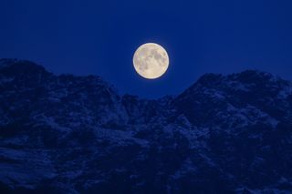 full moon rises behind snow-capped mountains.