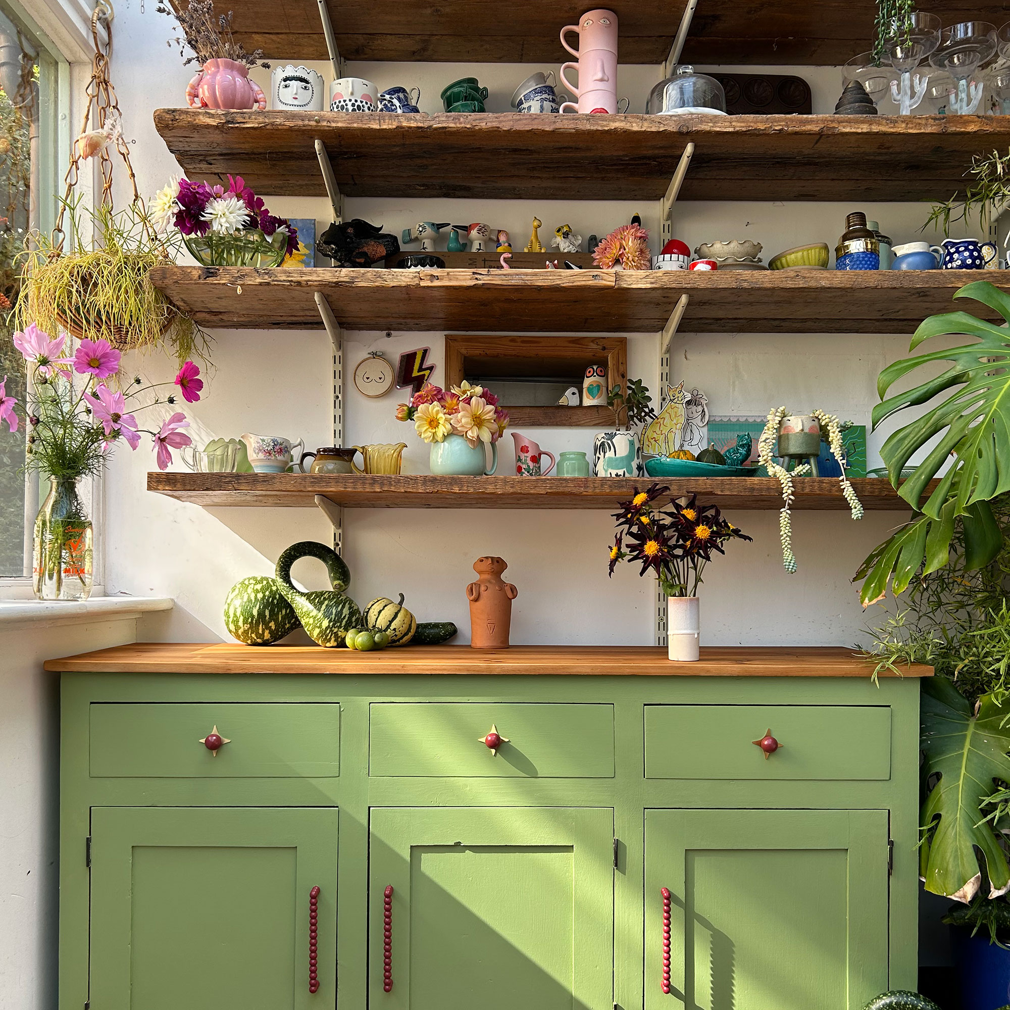 Green kitchen cabinets below open wooden shelving full of vegetables, mugs and vases of flowers