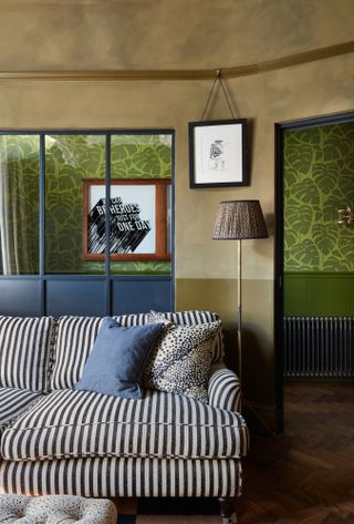 Living room with khaki two-tone limewash and matt painted walls, blue glazed partition, green patterned wallpaper in room to the rear, and striped black and white sofa