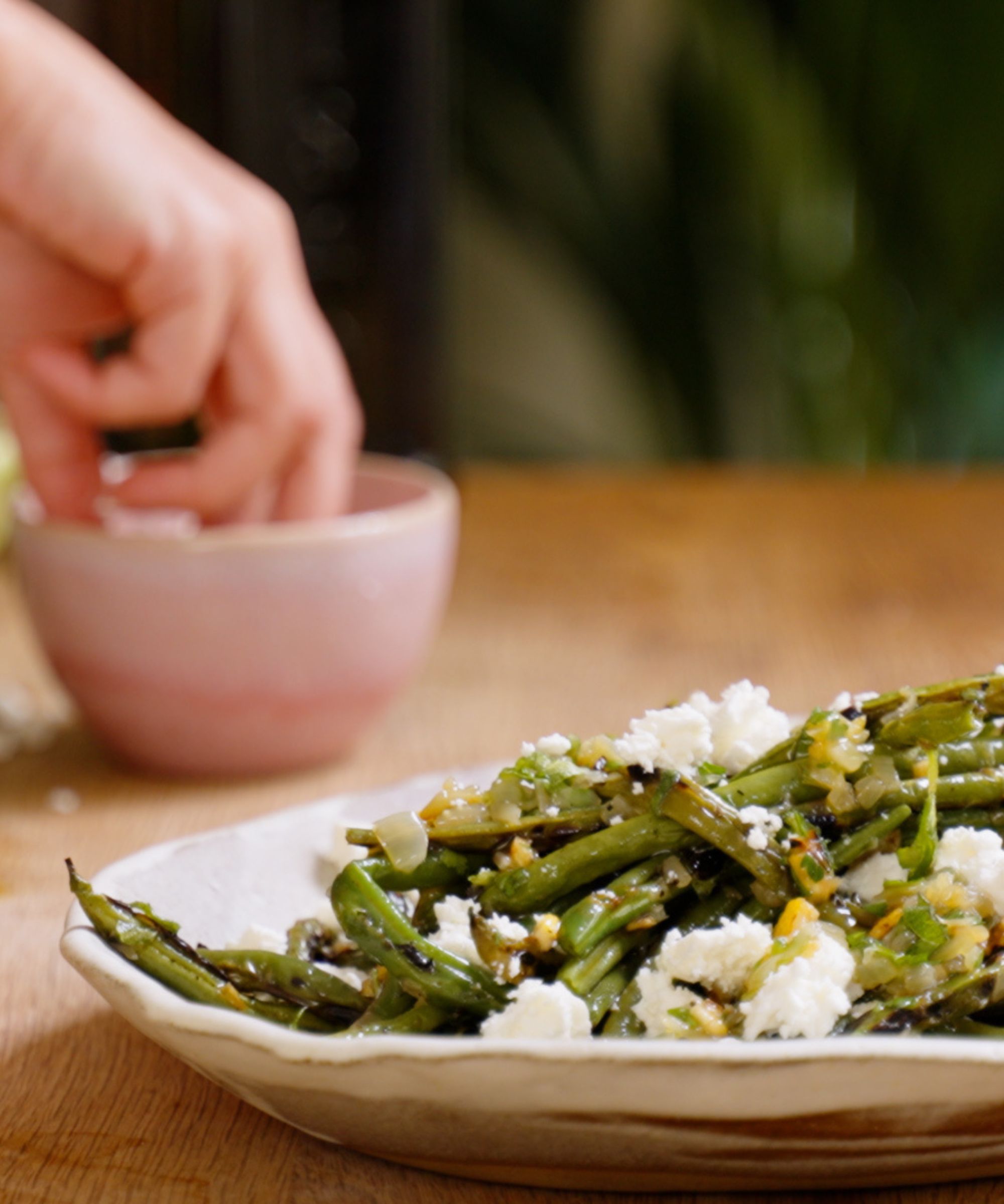 Plate of grilled green beans and feta with a hand in a pink pinch pot in the background